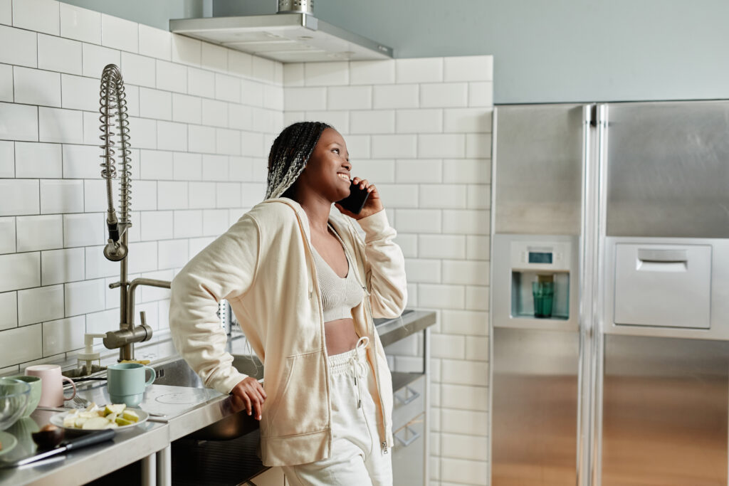 Side view portrait of young African-American woman speaking by smartphone while standing in kitchen interior