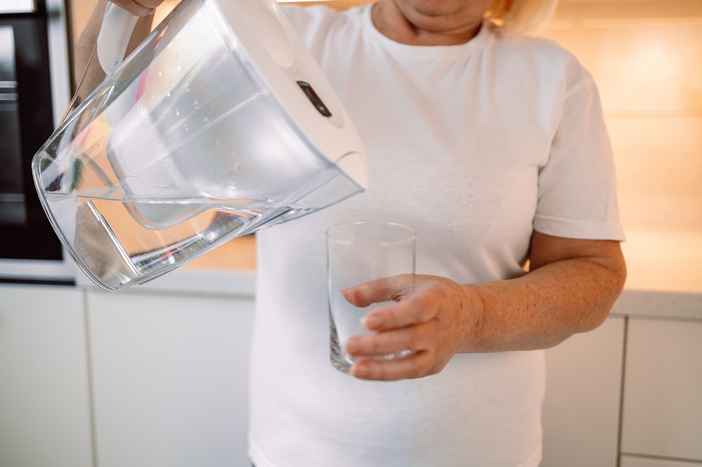 Smiling mature 60s woman pouring filtered water into glass at kitchen. Hydration, healthy lifestyle cocnept. Smiling senior woman pouring filtered water into glass, kitchen interior. Attractive older woman in casual outfit holding glass of fresh water, copy space. High quality photo